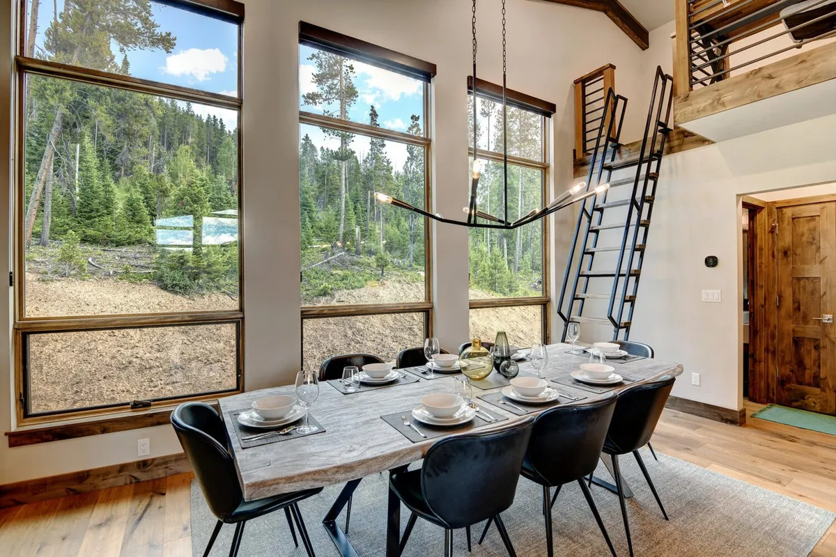 A welcoming dining area featuring a custom dining table positioned beside expansive windows that capture sunset hues and majestic landscapes. A ladder leads to a loft, and the room is set up for family dinners and friendly gatherings, exuding warmth and hospitality.