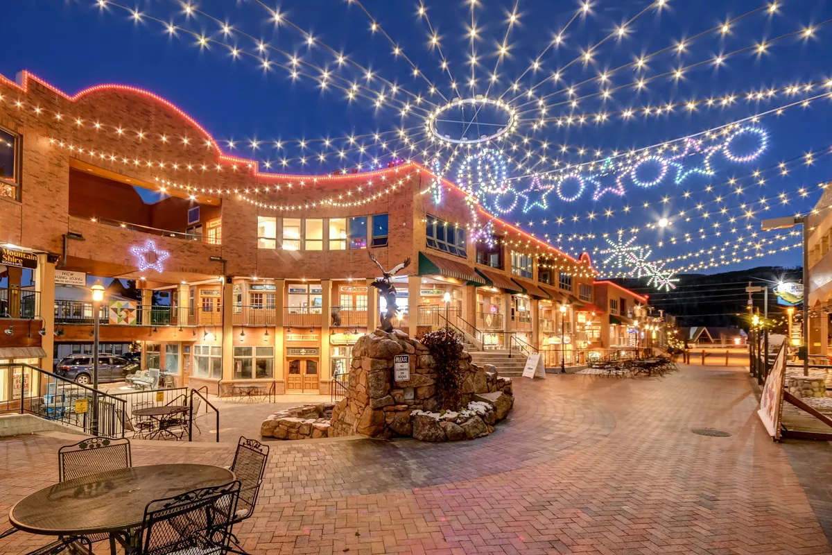 A lively exterior shot showing a plaza with decorative lights, a stone fountain, multiple seating areas, and storefronts. The atmosphere is festive, suggesting a hub for community events, shopping, dining, and outdoor gatherings.