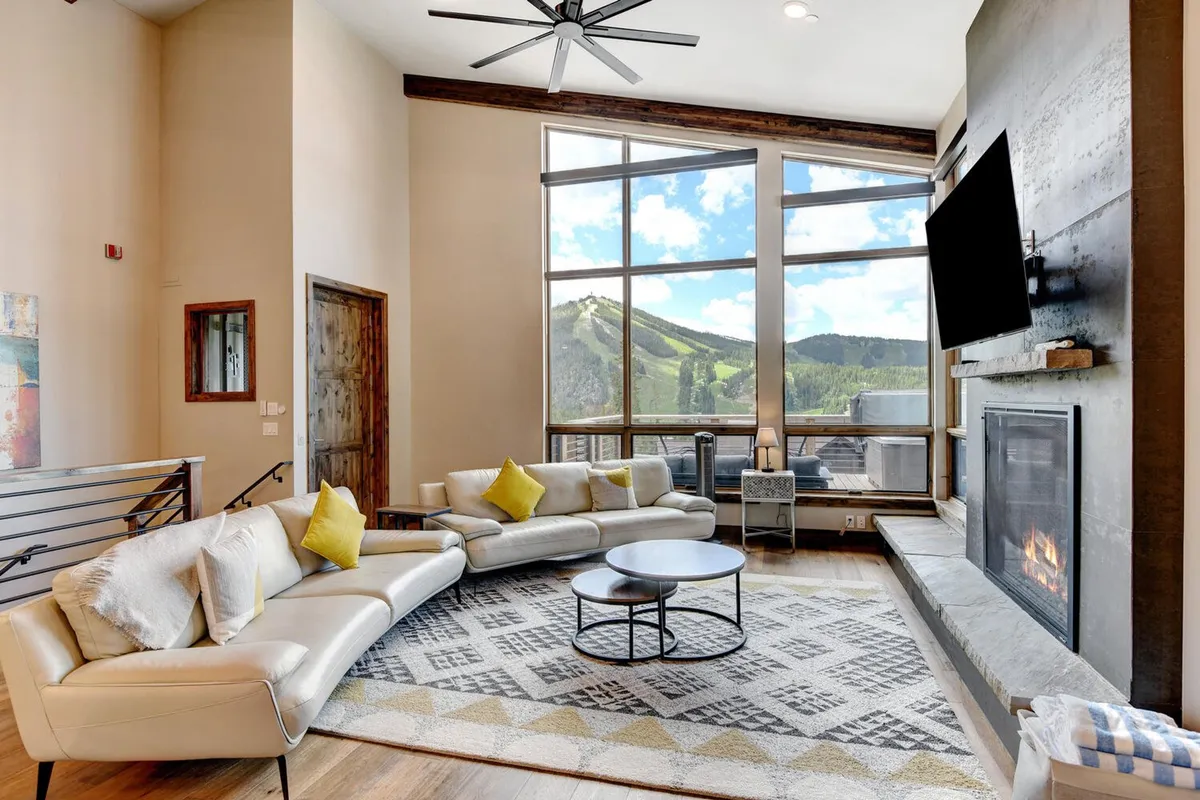 A spacious living room featuring floor-to-ceiling windows that showcase jaw-dropping mountain views. The room is centered around a stylish gas fireplace, with a comfortable curved sectional sofa and a modern coffee table inviting group relaxation or conversation. The abundant natural light, cozy textures, and striking vistas create an inviting, elevated space.