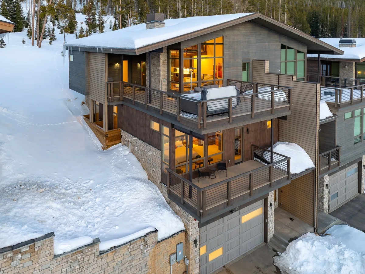 The image showcases the exterior of a luxury mountain villa in Winter Park, Colorado, featuring a snow-covered balcony, expansive stone and wood architecture, large windows glowing with warm light, and breathtaking views of snow-capped peaks at sunset. The inviting setting hints at après-ski relaxation and memorable gatherings, with the backdrop of pristine winter scenery.