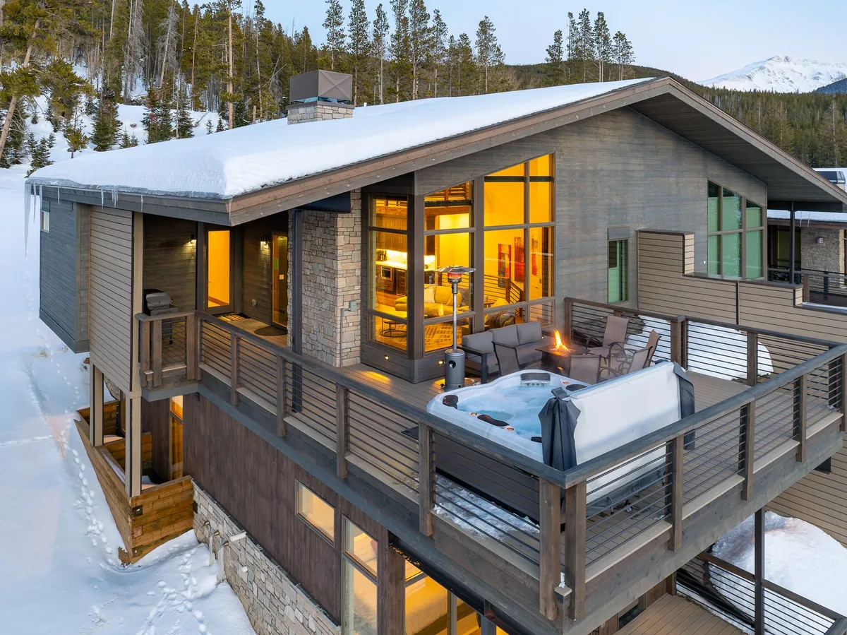 The image shows a hot tub on a snow-framed deck with an inviting outdoor seating area and large windows, all set against a backdrop of winter mountain scenery. The space looks luxurious and perfect for winter evenings with the contrast of warmth and snowy outdoors.