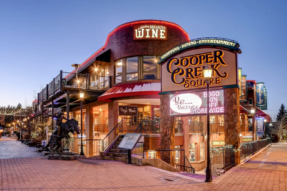 A lively exterior shot of the Cooper Creek Square sign surrounded by a wine shop and decorative lights, suggesting a bustling center for shopping, dining, and community events. The atmosphere is festive, hinting at local summer festivals and nightlife.