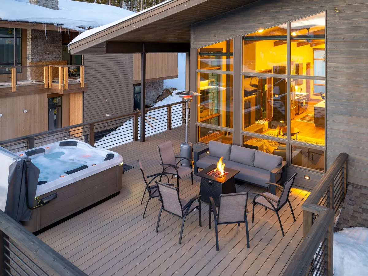 An exterior shot of a private outdoor seating area featuring a steaming hot tub, crackling fire pit, and comfortable chairs, all framed by large windows and gentle evening light. Snow blankets the ground, suggesting a perfect après-ski retreat.