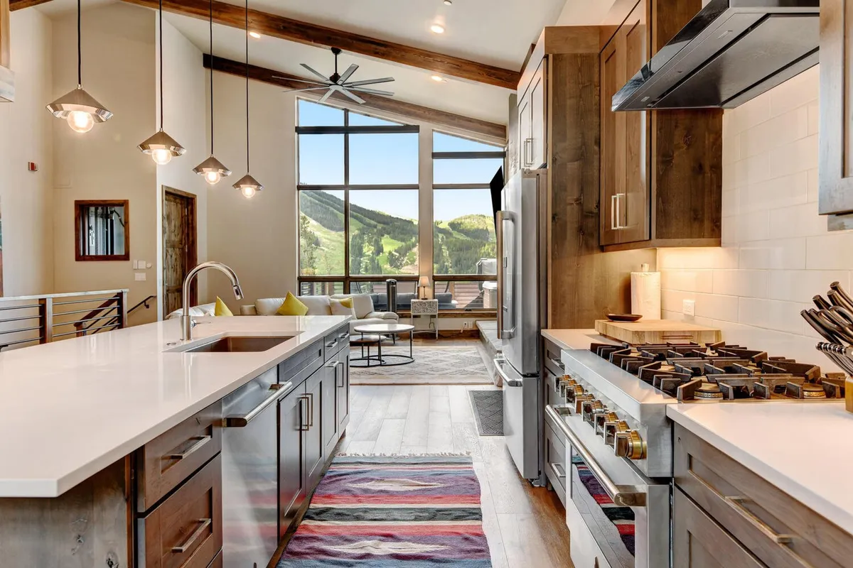 An open-concept kitchen and living area anchored by a modern kitchen island with stainless steel appliances. Large windows frame panoramic mountain views, while a decorative rug adds warmth to the space. The layout is perfect for cooking, entertaining, and enjoying scenic surroundings.