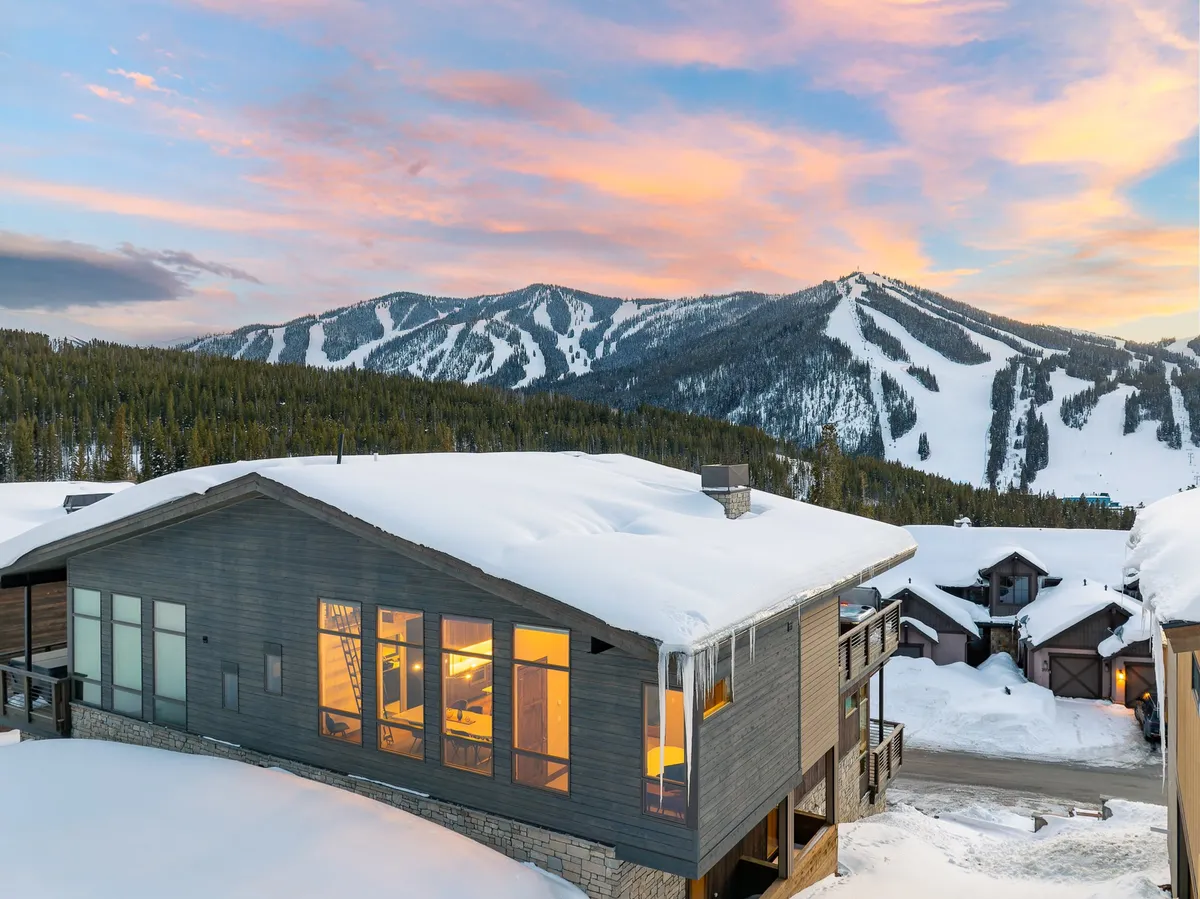 A glowing, cozy cabin set amid snow-covered mountains beneath a vibrant sunset sky. The warm interior lights suggest comfort and welcome, while the landscape alludes to nearby trails for skiing, snowboarding, and hiking. The porch offers a prime vantage for soaking up the natural beauty.