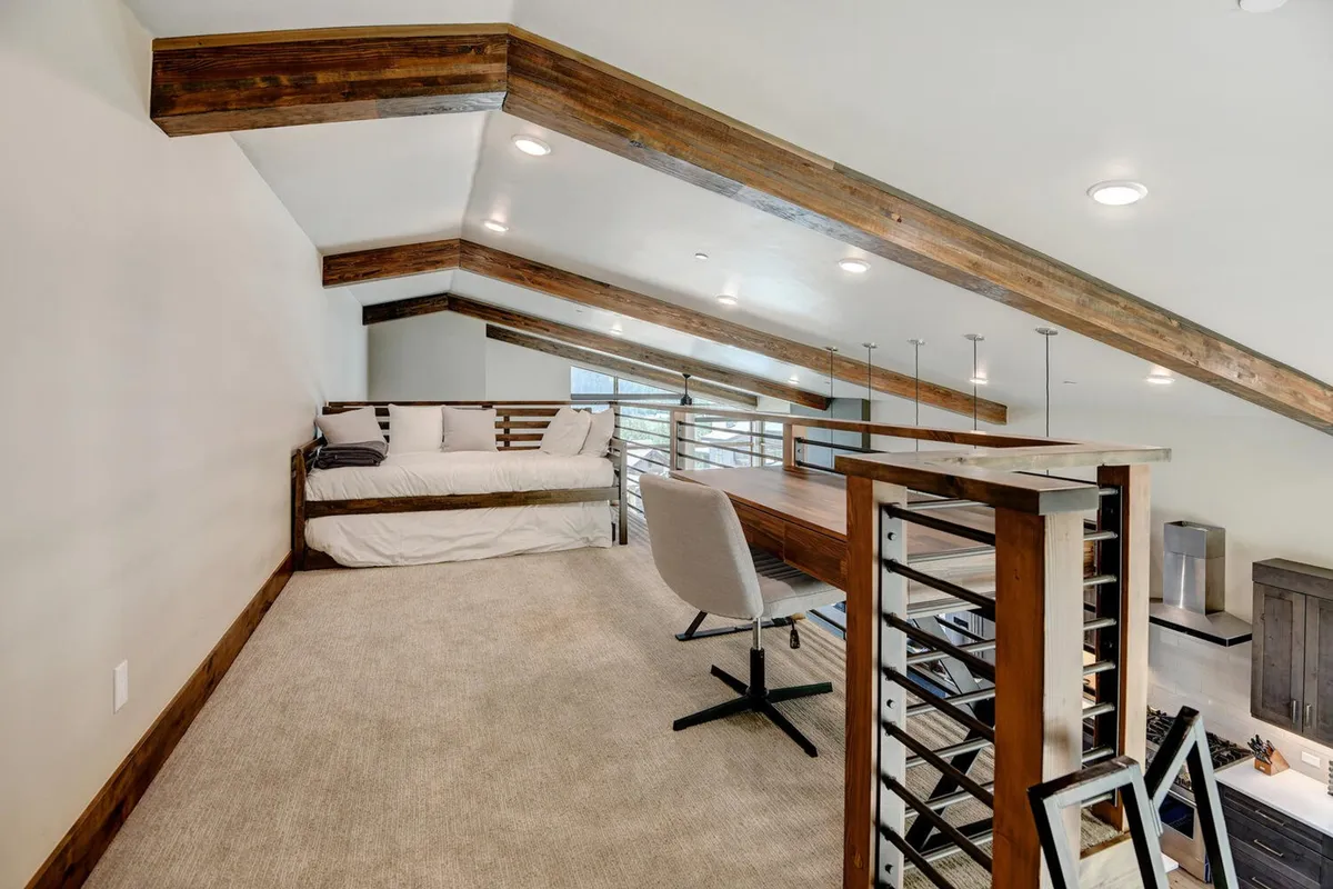 A lofted area showcasing a bed tucked into a cozy nook, a work desk with chair, exposed wooden beams, and plenty of natural light streaming through windows. The setting conveys both a restful and productive environment, ideal for kids or solo retreats.